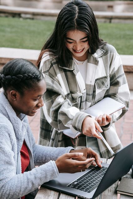 Two students exchanging information, one student has a pen and the other student a laptop.  They are sitting outside. 