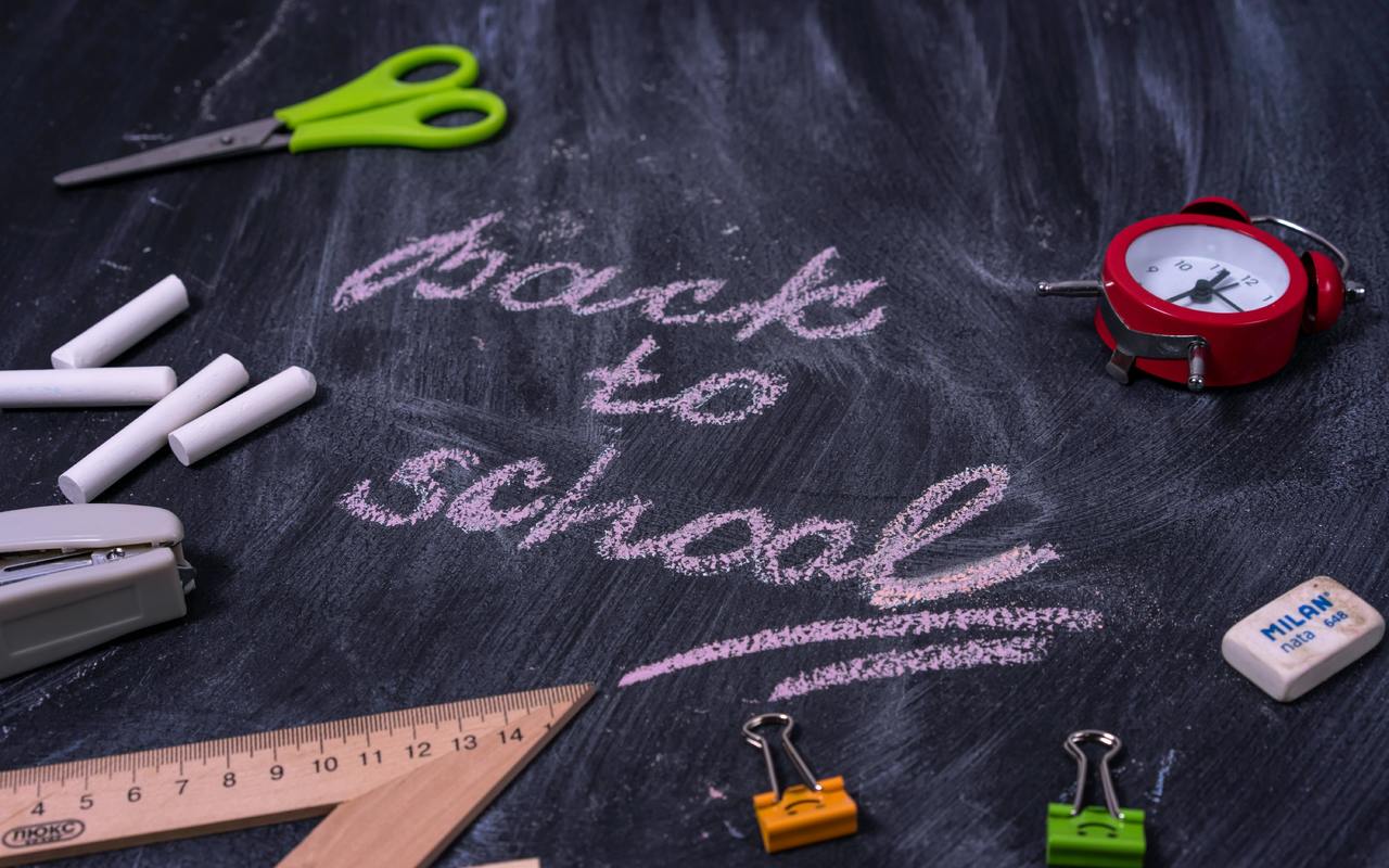 A photo of a chalk-smudged blackboard with the words "back to school" written in cursive and underlined twice. There are school supplies and other items scattered around the "back to school" message, which is in the center of the blackbaord. The supplies and items include these items: a pair of scissors, 4 pieces of chalk, an analog alarm clock, a stapler, a wooden triangular ruler, an eraser, and two binder clips.