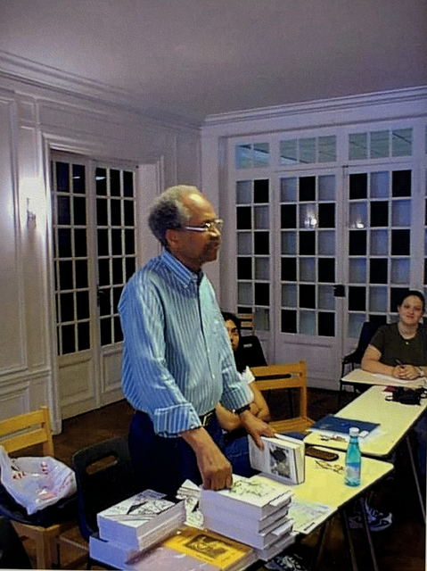 James A. Emanuel standing over a desk with one of his books in his hand.  He has a stack of books near him, which look to be 'Jazz from the Haiku King' and 'Whole Grain: Collected Poems 1958 to 1989.' Two students are looking at him. 