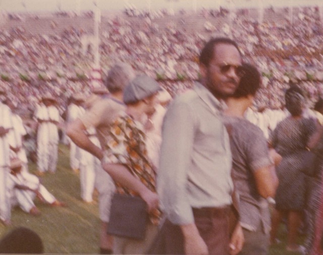 Emanuel stands in the midst of a crowd of people in stadium. He wears glasses. He stands to the side. He is facing the camera.