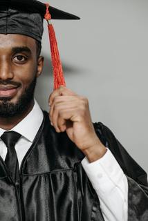 A close-up, vertically bifurcated portrait photograph of a young man. He is wearing a dress shirt and tie with a silky graduation robe layered on top. He is wearing a graduation cap and holding the brightly colored red tasel on his cap. Photo credits to Pavel Danilyuk on Pexels.com