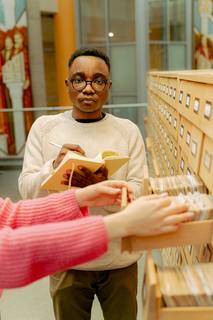 two hands that seem to be a librarian rummaging through an old-school card catalog in the foreground with a young man writing in a notepad and looking miserable in the background